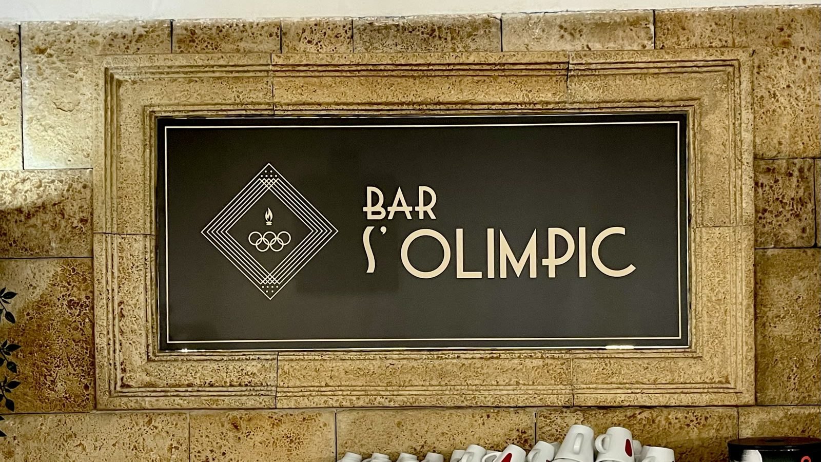 La placa esmaltada del Bar S'Olimpic darrere la barra: el rombe daurat de marcs concèntrics sobre fons fosc, amb el nom del bar al costat.